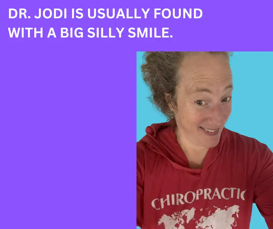 A person named Dr. Jodi is smiling in a red chiropractic hoodie with the text "DR. JODI IS USUALLY FOUND WITH A BIG SILLY SMILE" on a purple background.